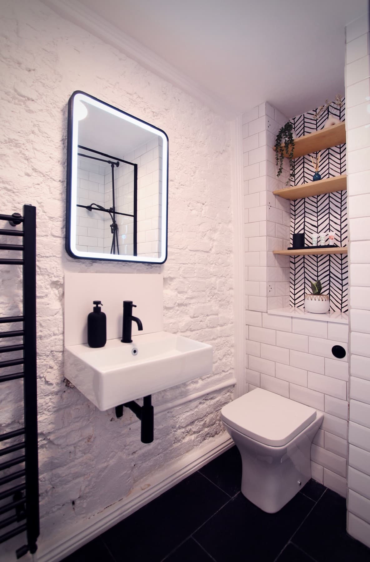 The bathroom — basin, lit mirror and black towel rail against painted brick